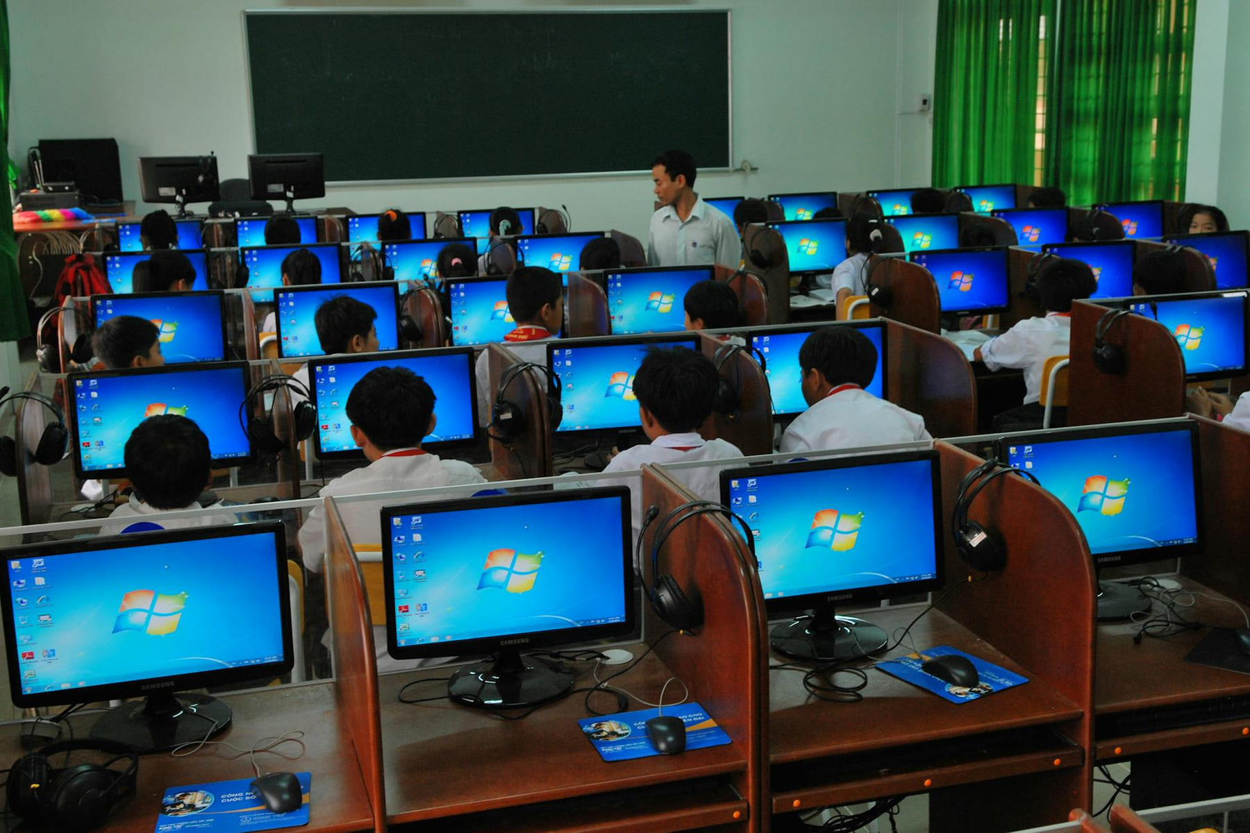 Students learning in a computer lab with multiple monitors and a teacher instructing. on The Course Compass